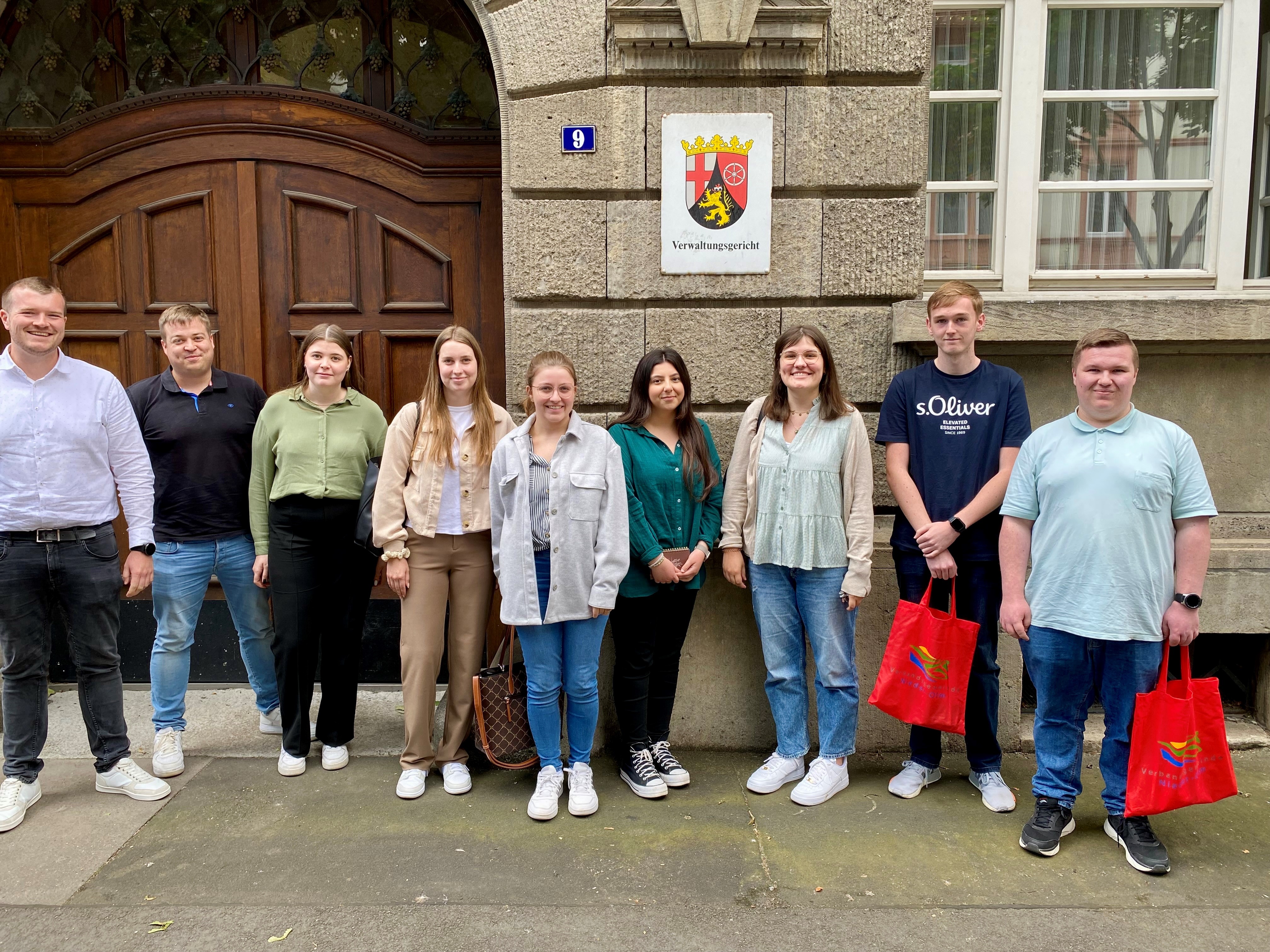 Auf dem Bild erkennt man eine Gruppe von circa zehn Personen. Diese stehen vor dem Verwaltungsgericht Mainz. Es sind die Auszubildenden der VG Nieder-Olm mit ihrem Ausbildungsleiter, der ganz links steht. Alle Personen haben den Blick nach vorne in Richtung des Betrachters.
