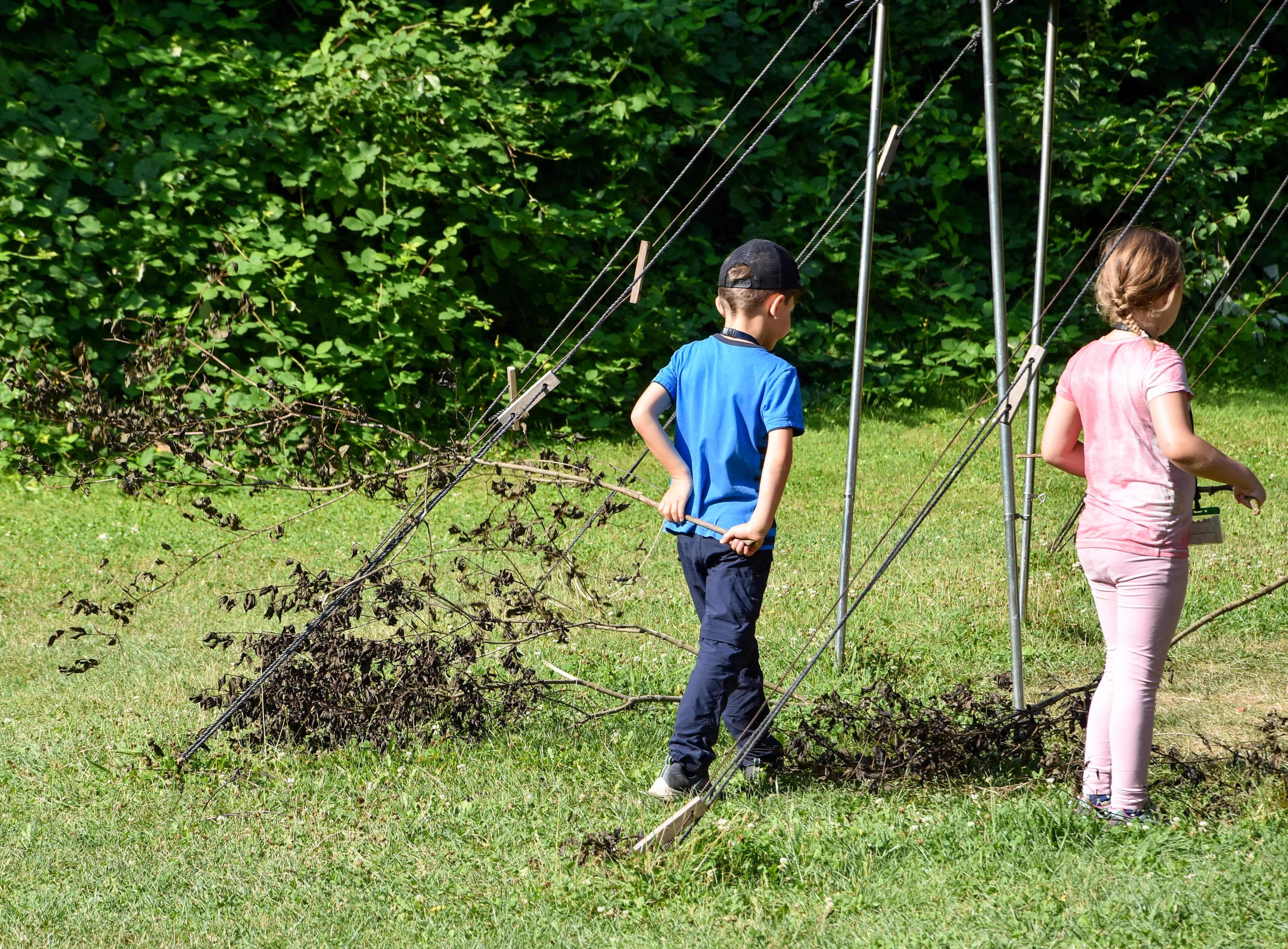 Zwei Kinder sind zu sehen. Diese stehen ind er grünen Natur und ziehen hinter sich einen großen Ast mit vertrockneten Blättern her. 