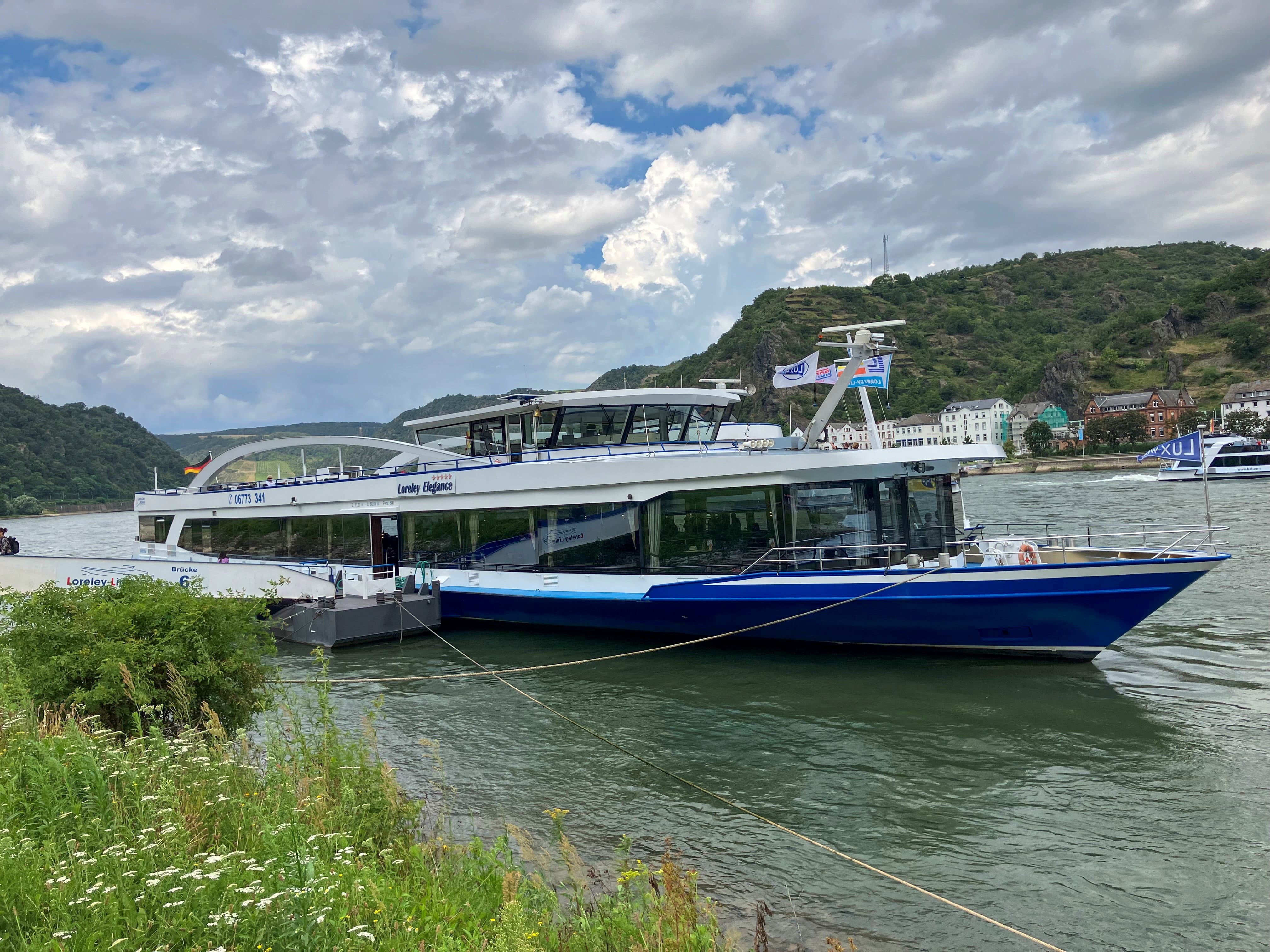 Auf dem Bild erkennt man ein Schiff. Dieses liegt an einem grünen Rhein-Ufer. Das Schiff ist blau und weiß. Am Himmel erkennt man weiße Wolken.