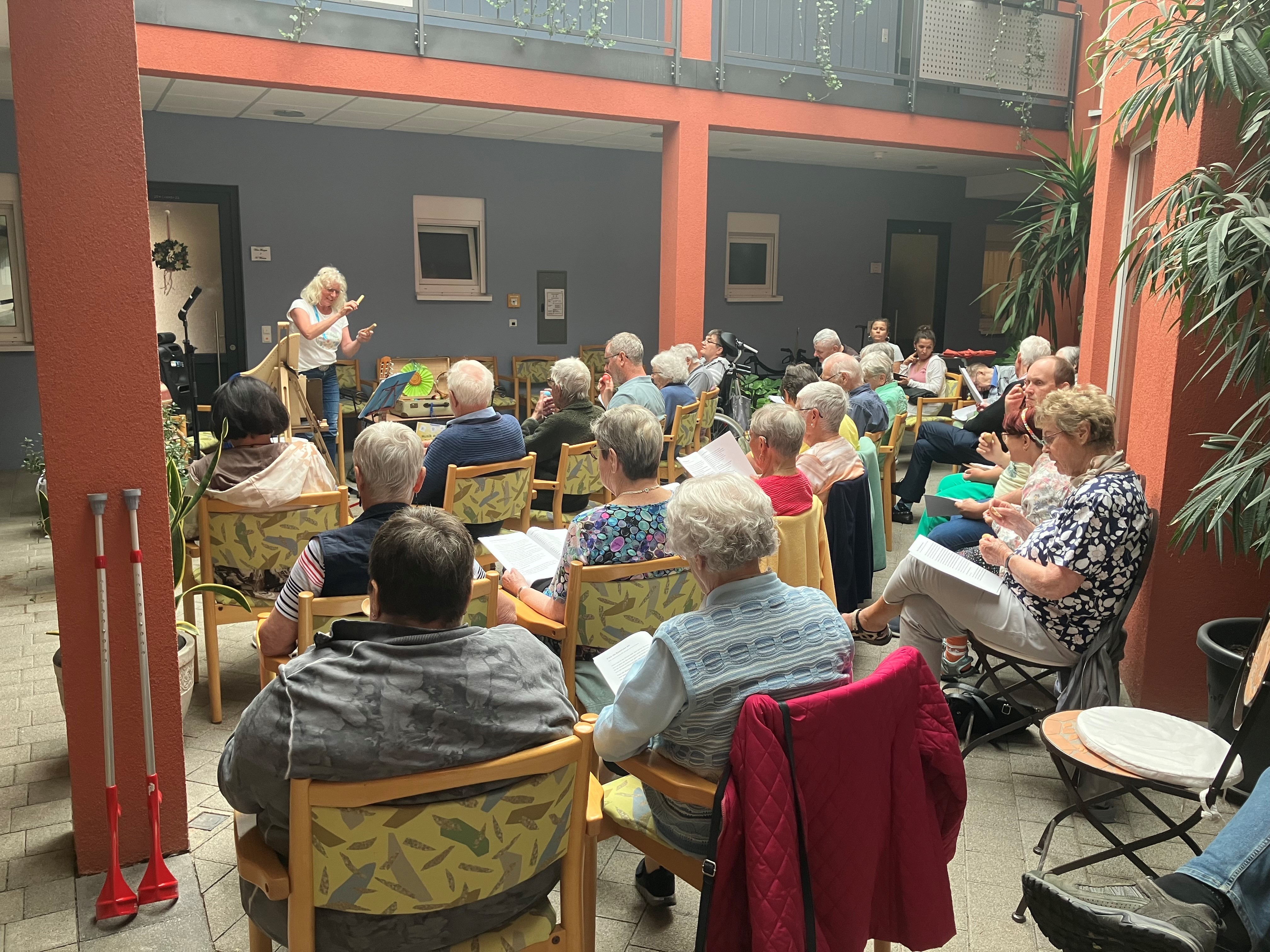 Auf dem Bild erkennt man eine Gruppe von Menschen, die auf Stühlen in einem halbrunden Kreis sitzen. Vorne steht eine Dame, die in ihrer Hand ein Musikinstrument hält. Die Gruppe der Menschen ist eine ältere Generation.