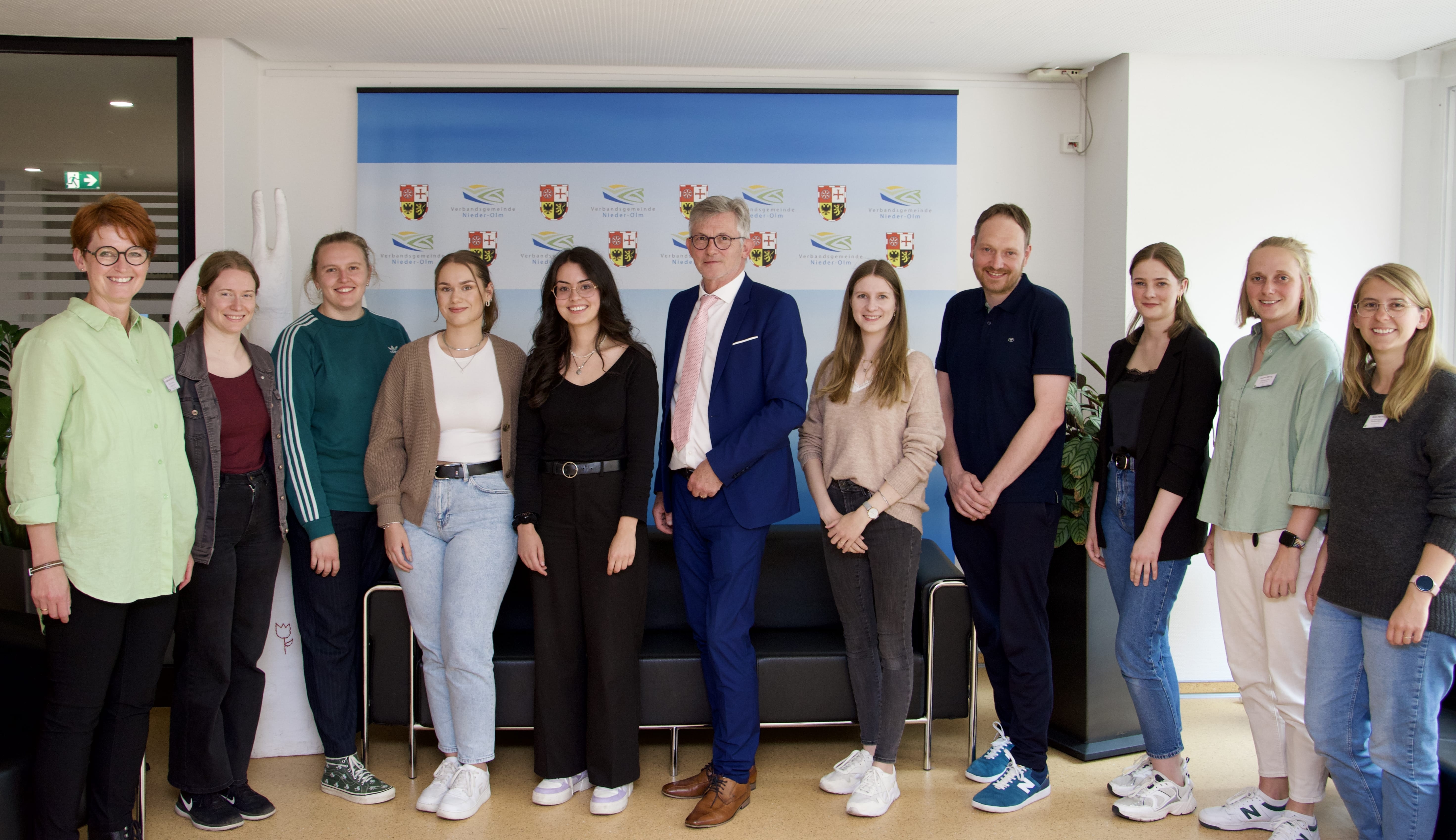 Auf dem Bild erkennt man eine Gruppe von circa zehn Personen. Diese stehen vor einer bunten Leinwand mit Logos und lachen in Richtung des Betrachters. In der Mitte ist der Bürgermeister der VG zu sehen. Er trägt einen dunkelblauen Anzug.