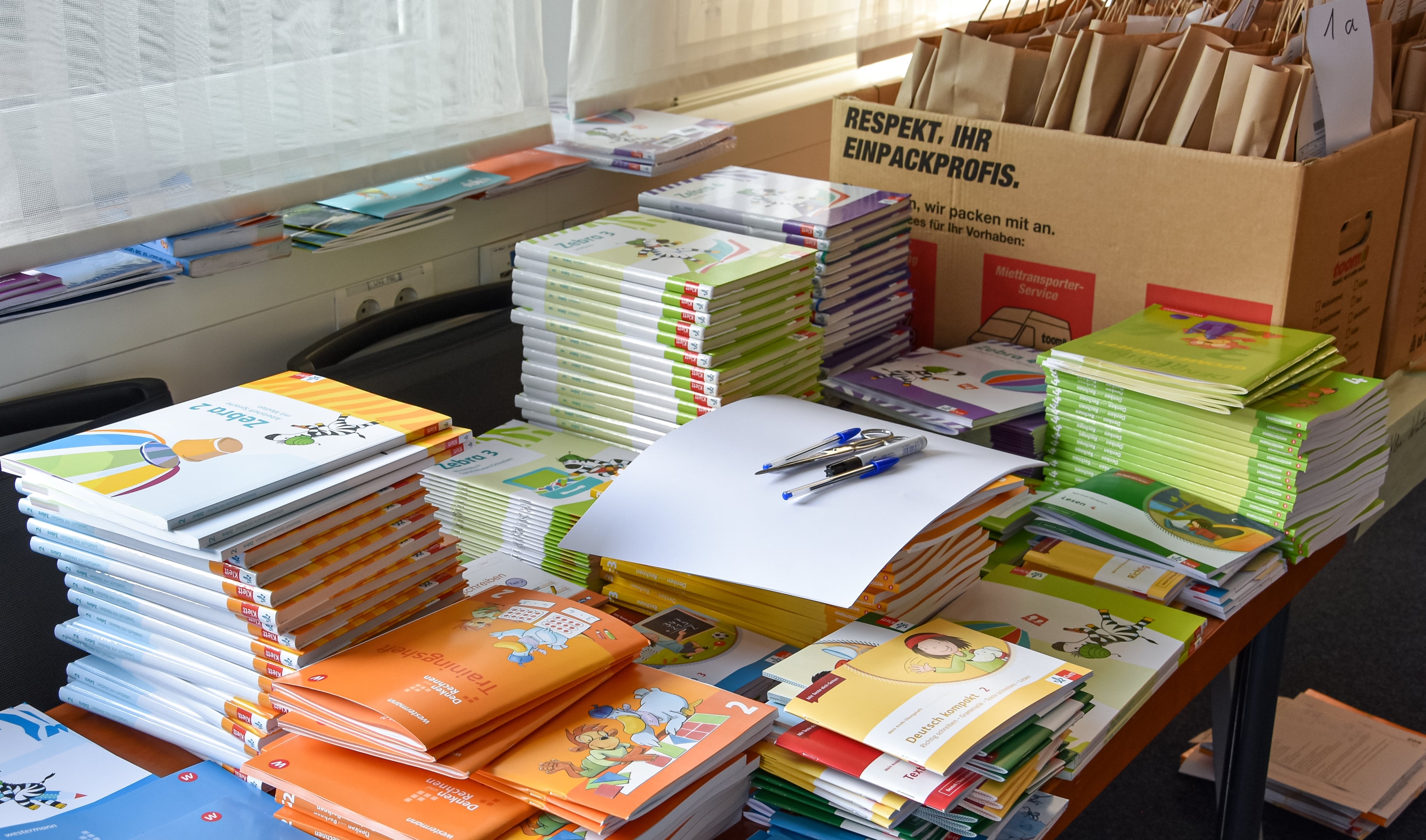 Schulbuchausleihe VG Nieder-Olm Auf dem Bild erkennt man diverse Schulbücher die auf verschiedenen Stapeln liegen.