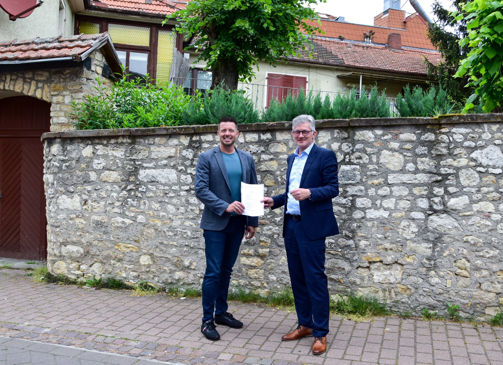 Ralph Spiegler und Dirk Hasenfuss stehen vor dem Gebäude in der Pfarrgasse. Man erkennt eine Backsteinmauer, sowie links ein altes, hohes Holztor. Die beiden Männer schauen fröhlich in die Kamera, halten ein Dokument in ihren Händen und tragen Anzüge. Es ist sonnig und im Hintergrund stehen grüne Laubbäume.