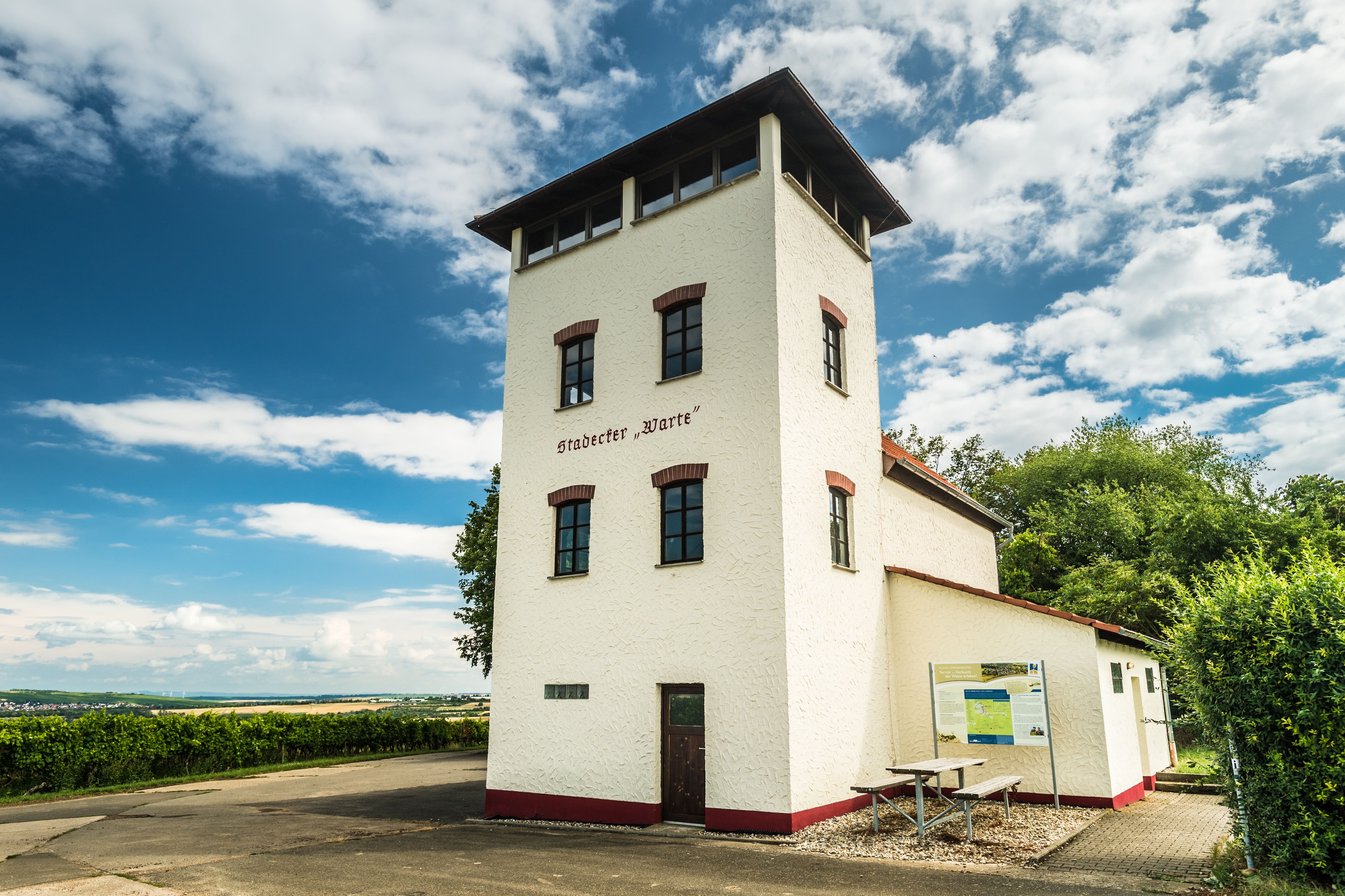 Auf dem Bild erkennt man ein hohes, altes Gebäude. Auf diesem steht in großen Lettern"" Stadecker Warte" geschrieben. Im Hintergrund erkennt man Weinberge sowie einen hellblauen Himmel. Es ist ein sommerlicher Tag,