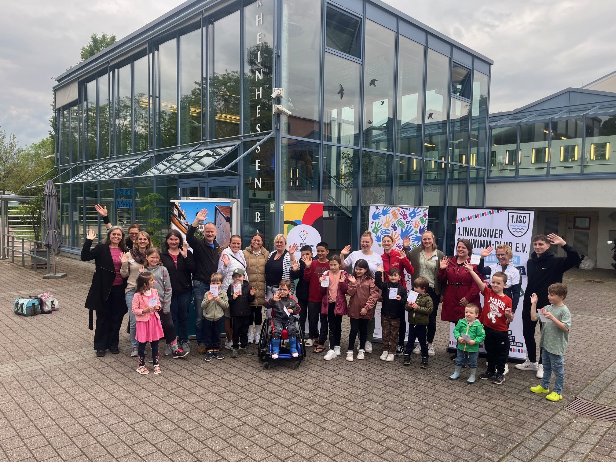 Auf dem Foto steht eine Gruppe von circa 20 Personen vor diversen Roll-Ups. Im Hintergrund erkennt man das Rheinhessen-Bad Nieder-Olm. Es sind die Teilnehmer und Trainer des inklusiven Schwimmkurses. So sieht man unter anderem zentral einen Jungen, der im Rollstuhl sitzt. 