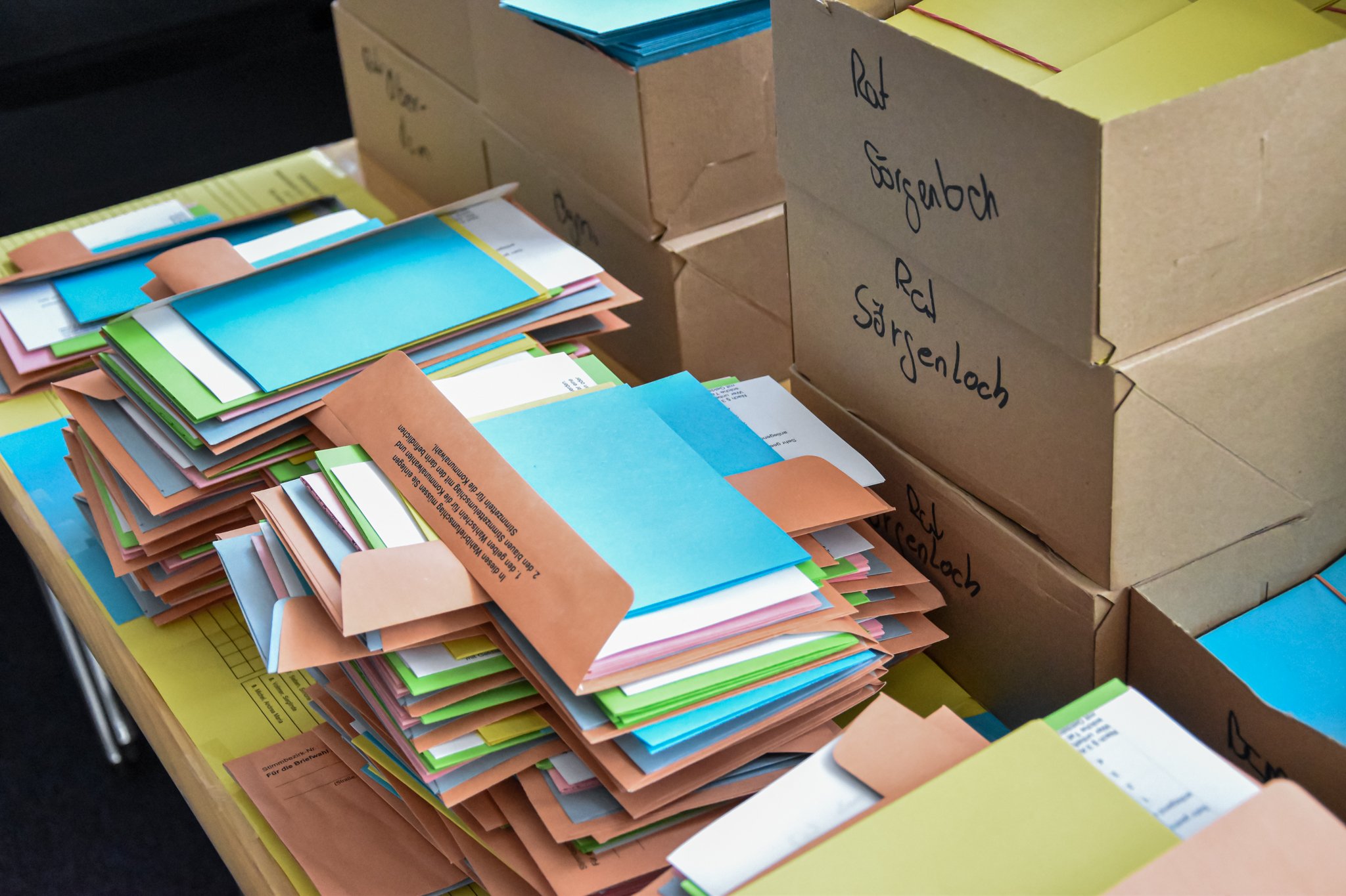 Arbeiten im Wahölbüro der Verbandsgemeinde Nieder-Olm Auf dem Foto sind zwei Stapel voller bunter Wahlbriefe und -Zettel zu erkennne. Man erkennt unter anderem die Farben Blau, Orange, Gelb und Grün. Im Hintergrund stehen diverse Kartons. Auf diesen sind die Ortsgemeinden der VG Nieder-Olm geschrieben.