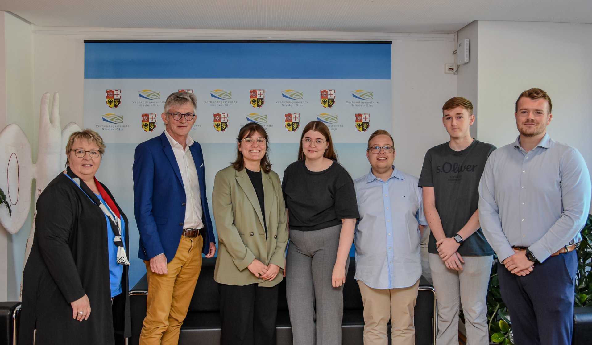 Auf dem Bild erkennt man eine Gruppe von sieben Personen, die vor einer Fotowand mit blauem Hintergrund stehen.