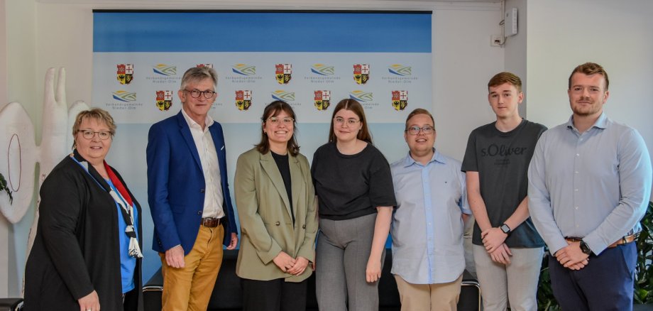 Auf dem Bild erkennt man eine Gruppe von sieben Personen, die vor einer Fotowand mit blauem Hintergrund stehen.