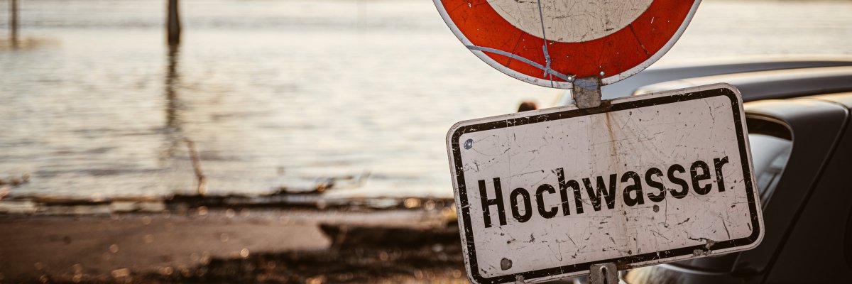 Hochwasser Auf dem Bild erkennt man im Hintergrund eine Menge Wasser. Im Vordergrund steht ein Schild mit der Unterschrift "Hochwasser"
