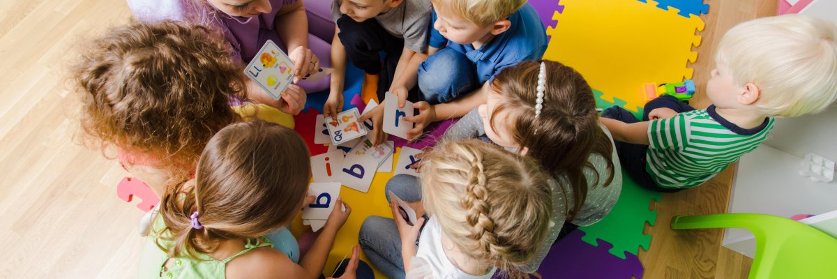 Kita Auf dem Bild erkennt man eine Erzieherin die auf bunten Kissen sitzt. In einem Kreis sitzen viele Kinder um sie herum. In ihren Händen hält die Erzieherin Buchstabenkarten. Das Szenario spielt sich in einer Kita ab.