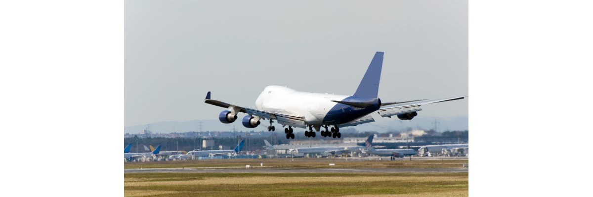 Auf dem Bild erkennt man ein weißes großes Flugzeug bei der Landung. Im Hintergrund sind weitere Flugzeuge auf einem Flughafen bzw. Rollfeld zu erkennen.