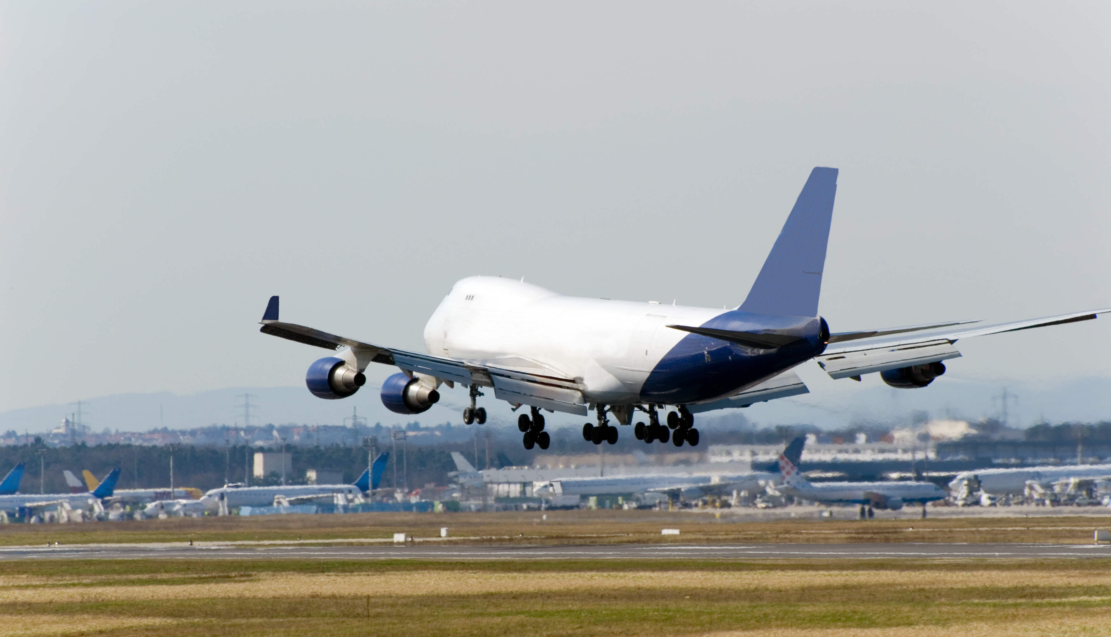 Flugzeug bei Landung Auf dem Bild erkennt man ein weißes großes Flugzeug bei der Landung. Im Hintergrund sind weitere Flugzeuge auf einem Flughafen bzw. Rollfeld zu erkennen.