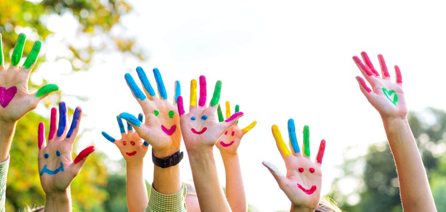 Auf dem Bild erkennt man Kinderhände. Diese sind bunt bemalt und man erkennt mehrere lachende Gesichter.