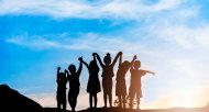 Die Silhouetten von Kindern sind zu sehen. Sie stehen auf einem Hügel, halten sich an den Händen und strecken die Arme nach oben. Im Hintergrund geht die Sonne unter.