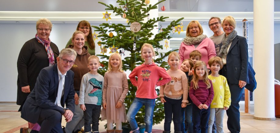 Gruppe von Erwachsenen und Kindern steht vor einem geschmückten Weihnachtsbaum in einem hellen Raum mit Säulen.