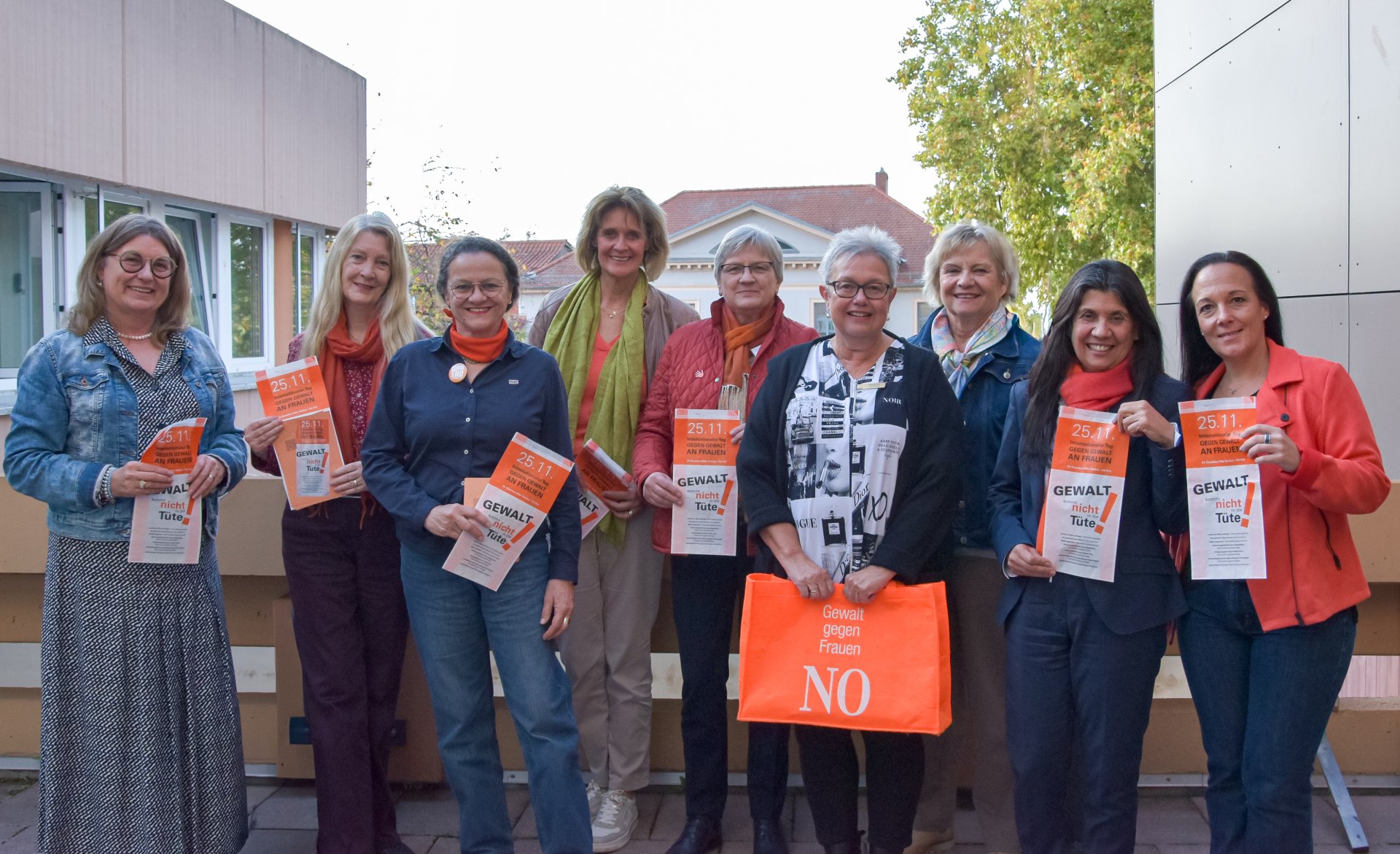 Auf diesem Foto erkennt man eine Gruppe von Frauen, die einen orangenen Flyer in die Kamera halten, auf einem Balkon stehen und in die Richtung des Betrachters blicken.