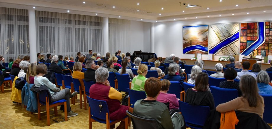 Mehrere Personen sitzen in einem Saal auf blauen Stühlen und hören einem Redner zu, der vor drei großen abstrakten Gemälden steht.