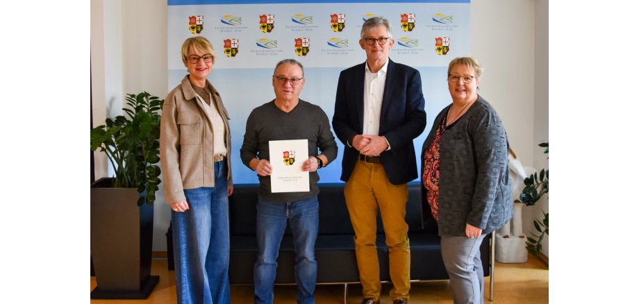 Auf dem Bild erkennt man vier Personen, die vor einer blauen Leinwand stehen und freudig in Richtung des Betrachtenden schauen.