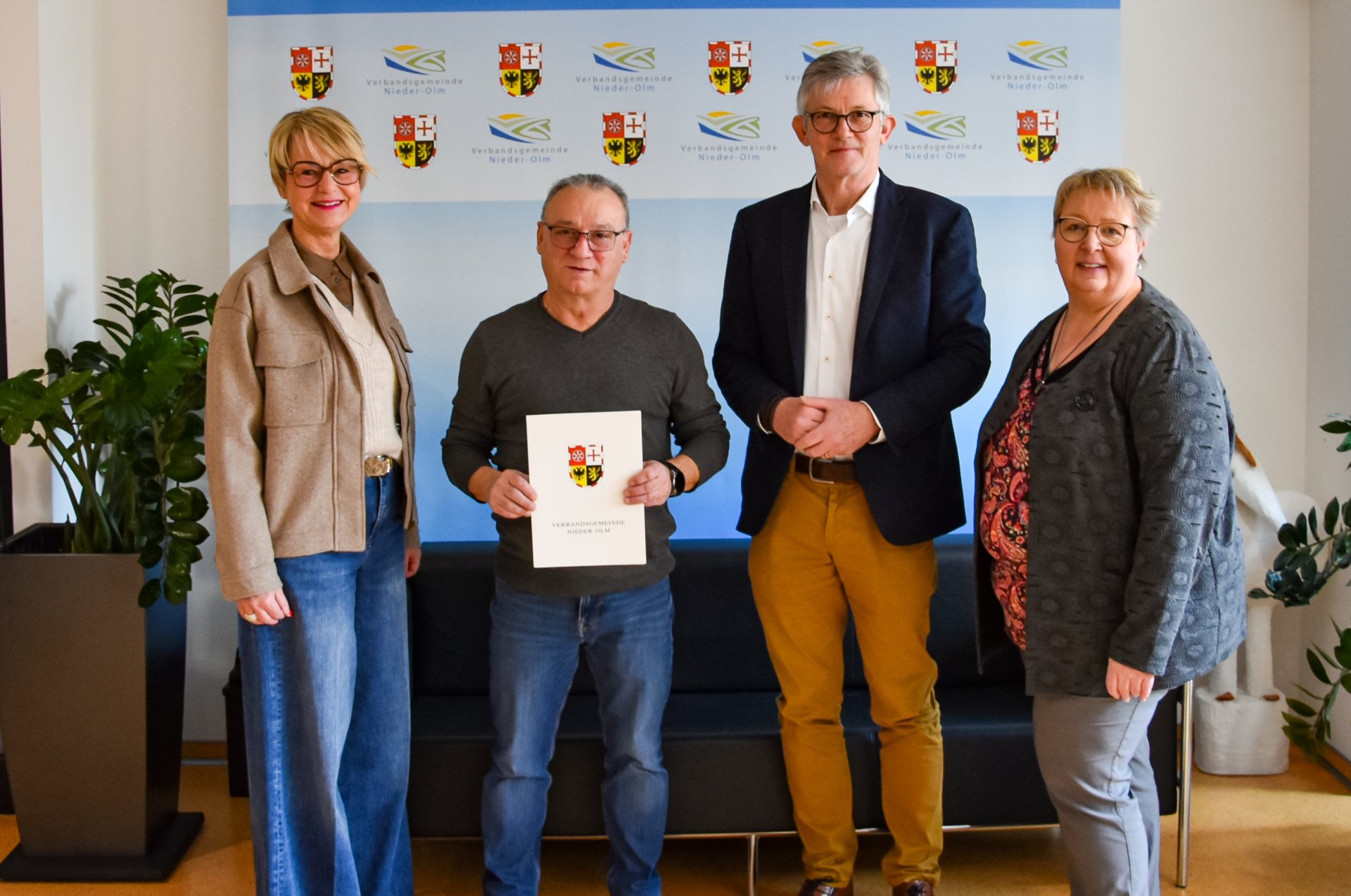 Auf dem Bild erkennt man vier Personen, die vor einer blauen Leinwand stehen und freudig in Richtung des Betrachtenden schauen.