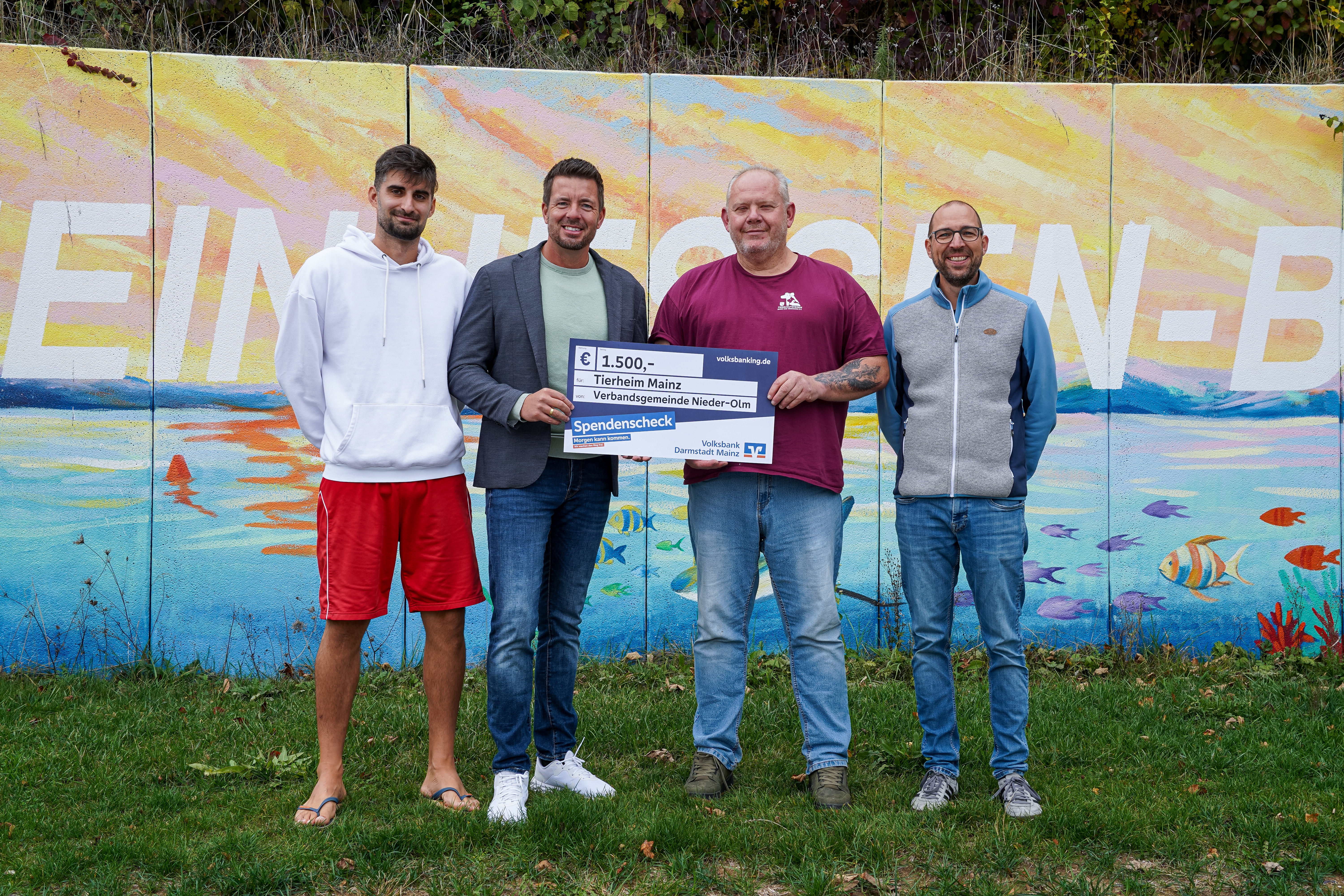 Ein Gruppenfoto von vier Personen vor einer bunten Wand im Rheinhessen-Bad NIeder-Olm. Es wird ein blauer Spendenscheck überreicht. 