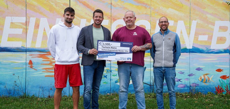Ein Gruppenfoto von vier Personen vor einer bunten Wand im Rheinhessen-Bad NIeder-Olm. Es wird ein blauer Spendenscheck überreicht. 