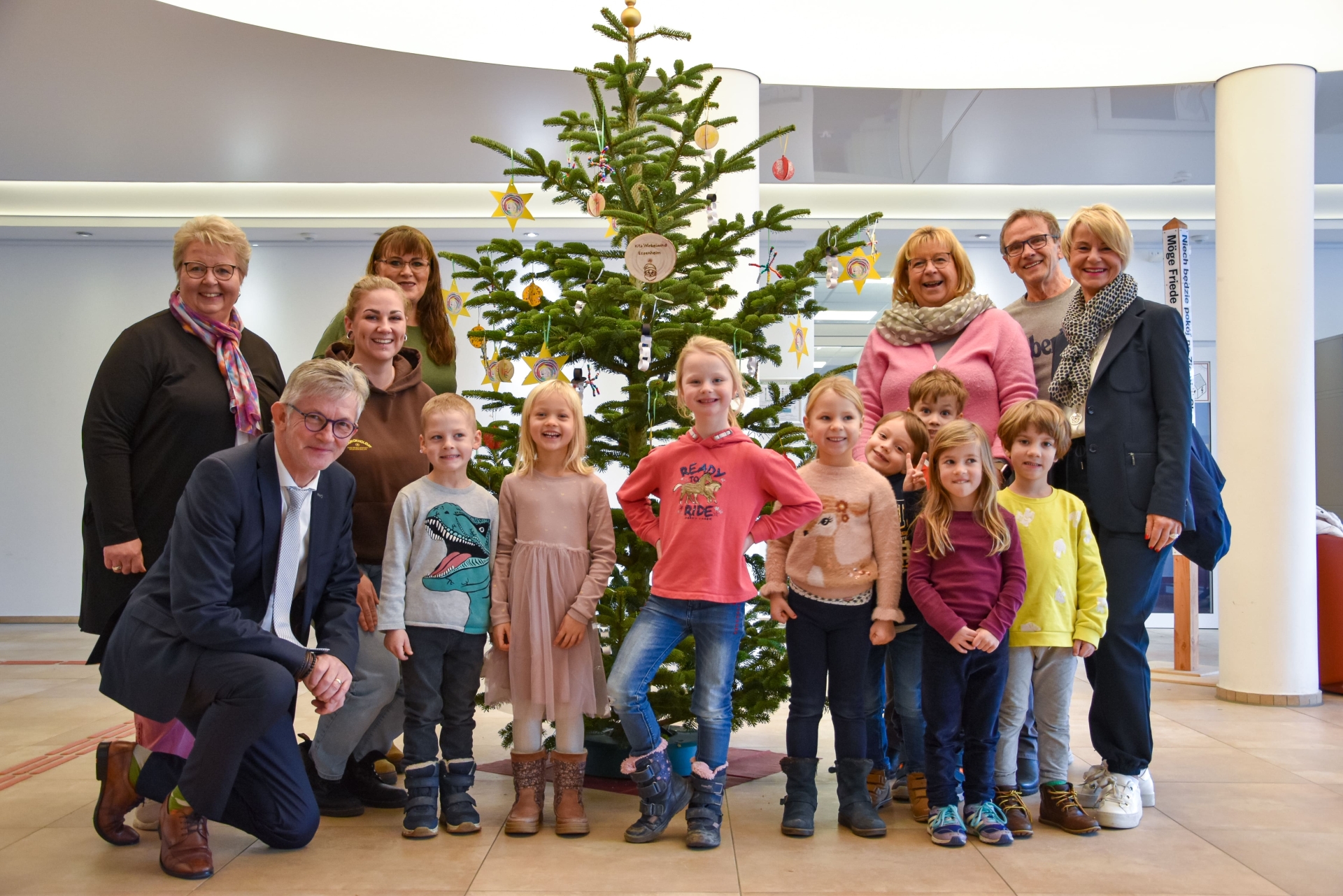 Gruppe von Erwachsenen und Kindern steht vor einem geschmückten Weihnachtsbaum in einem hellen Raum mit Säulen.