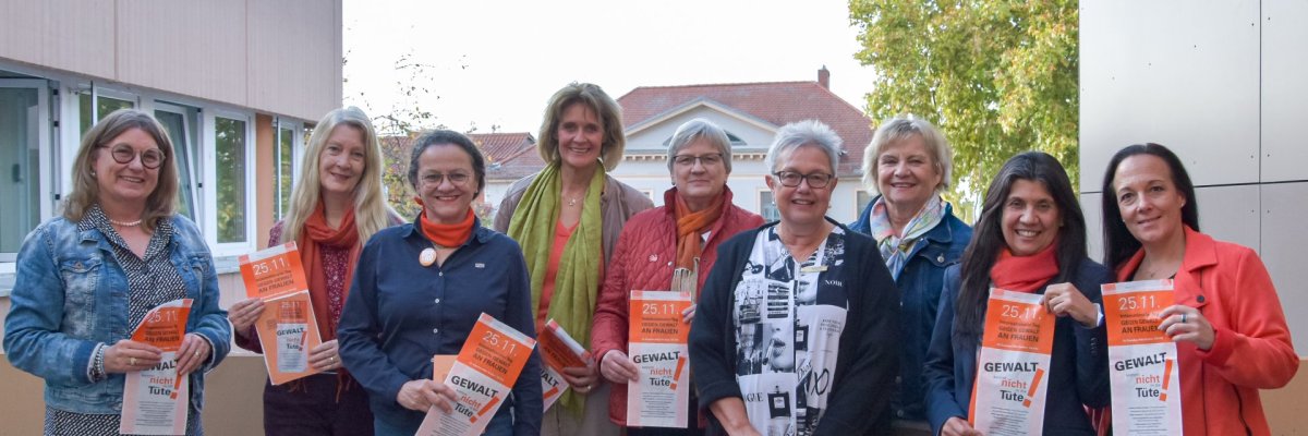 Auf diesem Foto erkennt man eine Gruppe von Frauen, die einen orangenen Flyer in die Kamera halten, auf einem Balkon stehen und in die Richtung des Betrachters blicken.