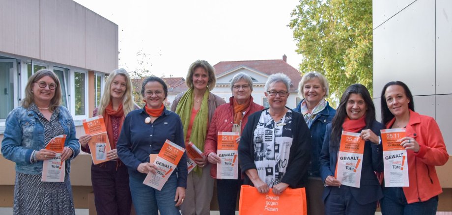 Auf diesem Foto erkennt man eine Gruppe von Frauen, die einen orangenen Flyer in die Kamera halten, auf einem Balkon stehen und in die Richtung des Betrachters blicken.
