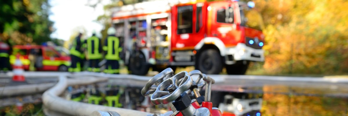 Auf dem Foto sieht man in Entfernung einen Feuerwehrwagen. Im Vordergrund liegen Schläuche.