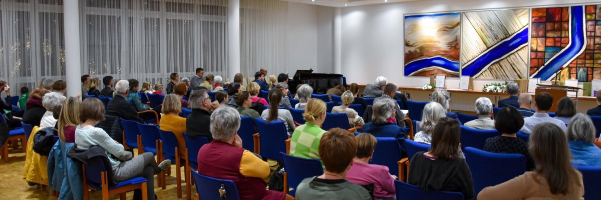 Mehrere Personen sitzen in einem Saal auf blauen Stühlen und hören einem Redner zu, der vor drei großen abstrakten Gemälden steht.
