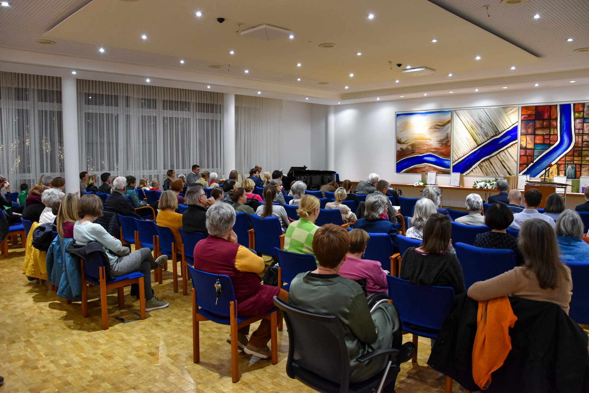 Mehrere Personen sitzen in einem Saal auf blauen Stühlen und hören einem Redner zu, der vor drei großen abstrakten Gemälden steht.